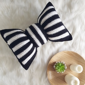 Puede incluir: Una almohada de crochet a rayas blancas y negras con forma de lazo. La almohada está sobre una superficie blanca y esponjosa.