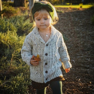 K&ouml;nnte beinhalten: Ein kleines Kind tr&auml;gt einen wei&szlig;en Strickcardigan mit einem gr&uuml;nen Schleifenhaarband, einen braunen Rock und dunkelgr&uuml;ne Strumpfhosen. Das Kind geht auf einem Weg mit braunem Mulch.