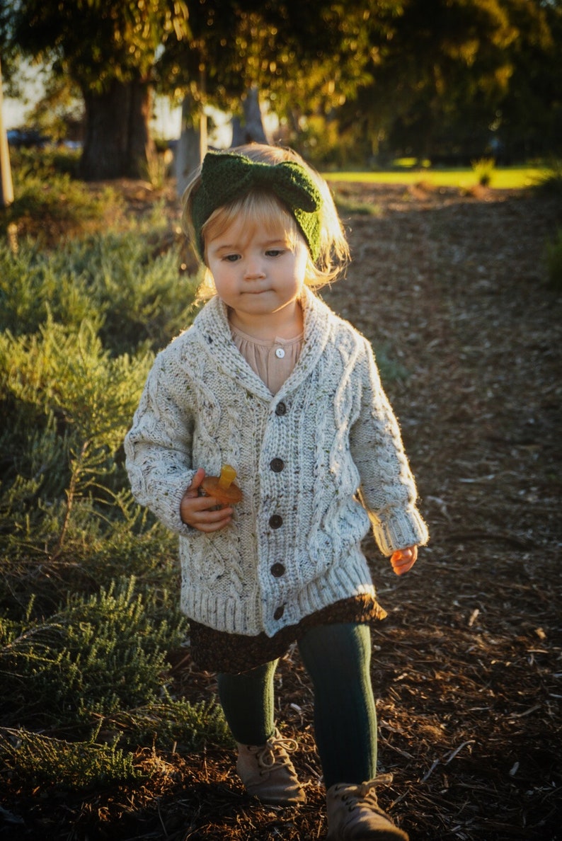 K&ouml;nnte beinhalten: Ein kleines Kind tr&auml;gt einen wei&szlig;en Strickcardigan mit einem gr&uuml;nen Schleifenhaarband, einen braunen Rock und dunkelgr&uuml;ne Strumpfhosen. Das Kind geht auf einem Weg mit braunem Mulch.