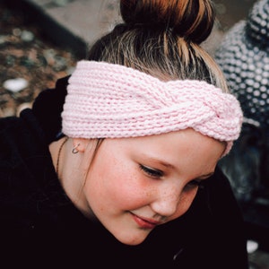 Può includere: Un cerchietto di lana rosa con un design intrecciato. Il cerchietto è indossato da una persona con i capelli castani.