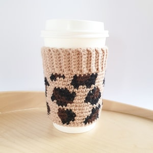May include: A beige and brown crochet coffee cup sleeve with a leopard print design. The sleeve is wrapped around a white disposable coffee cup with a lid. The sleeve is made of a soft, textured yarn.
