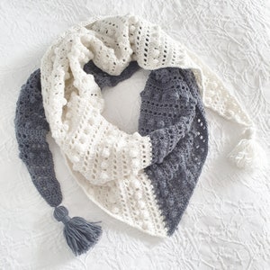 May include: A crocheted shawl in a white and grey colour scheme. The shawl has a textured design with bobbles and is finished with tassels.
