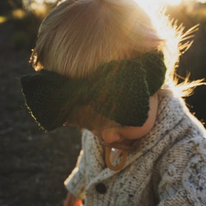 K&ouml;nnte beinhalten: Ein Kind tr&auml;gt einen wei&szlig;en Strickcardigan mit einem gr&uuml;nen Strickband als Kopfbedeckung. Das Kind steht auf einem Feld mit der Sonne, die hinter ihm scheint.