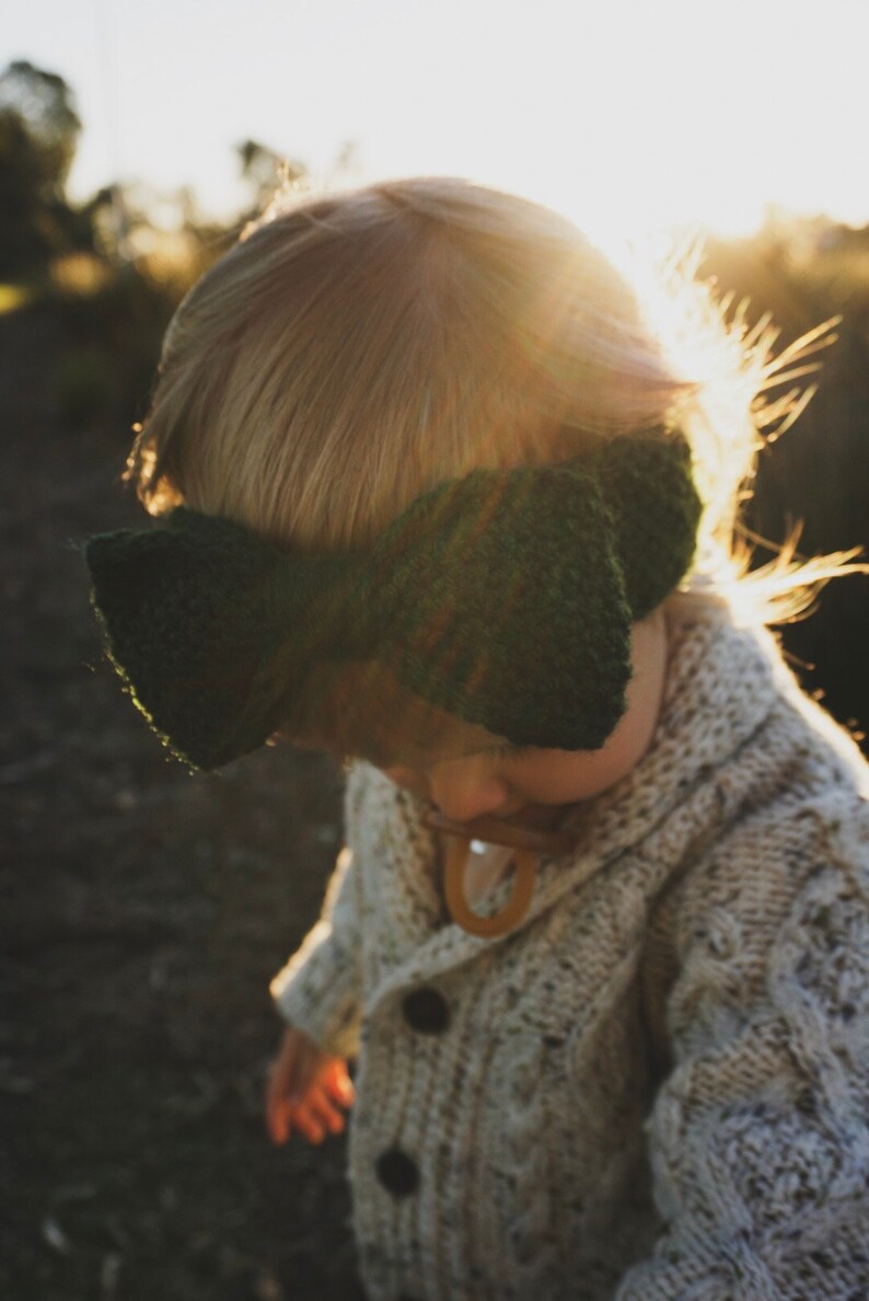 K&ouml;nnte beinhalten: Ein Kind tr&auml;gt einen wei&szlig;en Strickcardigan mit einem gr&uuml;nen Strickband als Kopfbedeckung. Das Kind steht auf einem Feld mit der Sonne, die hinter ihm scheint.