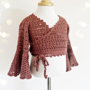 May include: A rust-coloured crocheted wrap top with bell sleeves and a tie closure. The top is made with a delicate open stitch pattern.