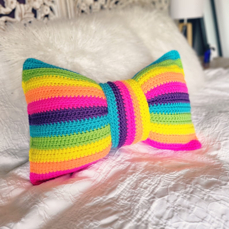 Pu&ograve; includere: Un cuscino a forma di fiocco in crochet a righe arcobaleno colorate. Il cuscino &egrave; realizzato con filato rosa acceso, arancione, giallo, verde, blu, viola e fucsia.