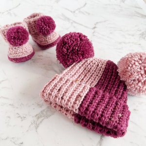 May include: A pink and burgundy knitted baby hat with two pom-poms. The hat is made of a soft, textured yarn.