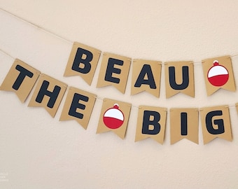 May include: A brown paper banner with black letters that reads "Beau is the big one" with red and white fishing bobbers between the words.