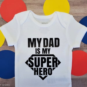 May include: White baby onesie with black text that reads "My Dad is My Super Hero" in a superhero logo design.