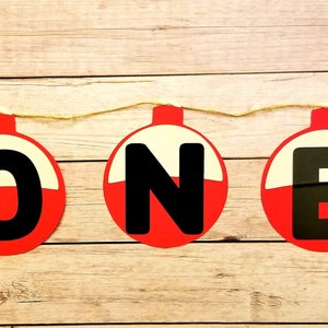 K&ouml;nnte beinhalten: Ein rotes, wei&szlig;es und schwarzes Angler-Schwimmer-Banner, das "ONE" buchstabiert. Das Banner ist mit einer Schnur befestigt und hat an beiden Enden blaue Fisch-Ausschnitte.