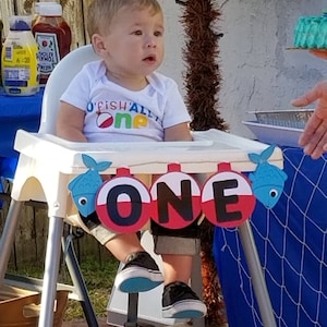 K&ouml;nnte beinhalten: Ein kleiner Junge in einem wei&szlig;en Strampler mit dem Text "O'Fish'ally One" sitzt in einem Hochstuhl. Vor dem Baby befindet sich ein Banner mit dem Wort "ONE" in schwarzen Buchstaben auf roten und wei&szlig;en Kreisen. Das Banner ist mit blauen Fischen verziert.