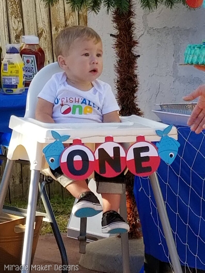 K&ouml;nnte beinhalten: Ein kleiner Junge in einem wei&szlig;en Strampler mit dem Text "O'Fish'ally One" sitzt in einem Hochstuhl. Vor dem Baby befindet sich ein Banner mit dem Wort "ONE" in schwarzen Buchstaben auf roten und wei&szlig;en Kreisen. Das Banner ist mit blauen Fischen verziert.