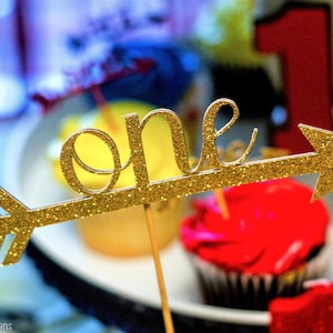 May include: Gold glitter arrow with the word "one" on it. The arrow is on a stick and is in front of cupcakes.