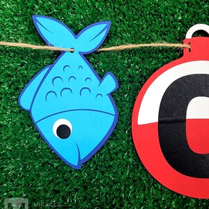 K&ouml;nnte beinhalten: Ein blauer Fisch und eine rot-wei&szlig;e Boje mit einem schwarzen Buchstaben C, die an einer Schnur h&auml;ngen. Der Hintergrund ist gr&uuml;nes Kunstgras.