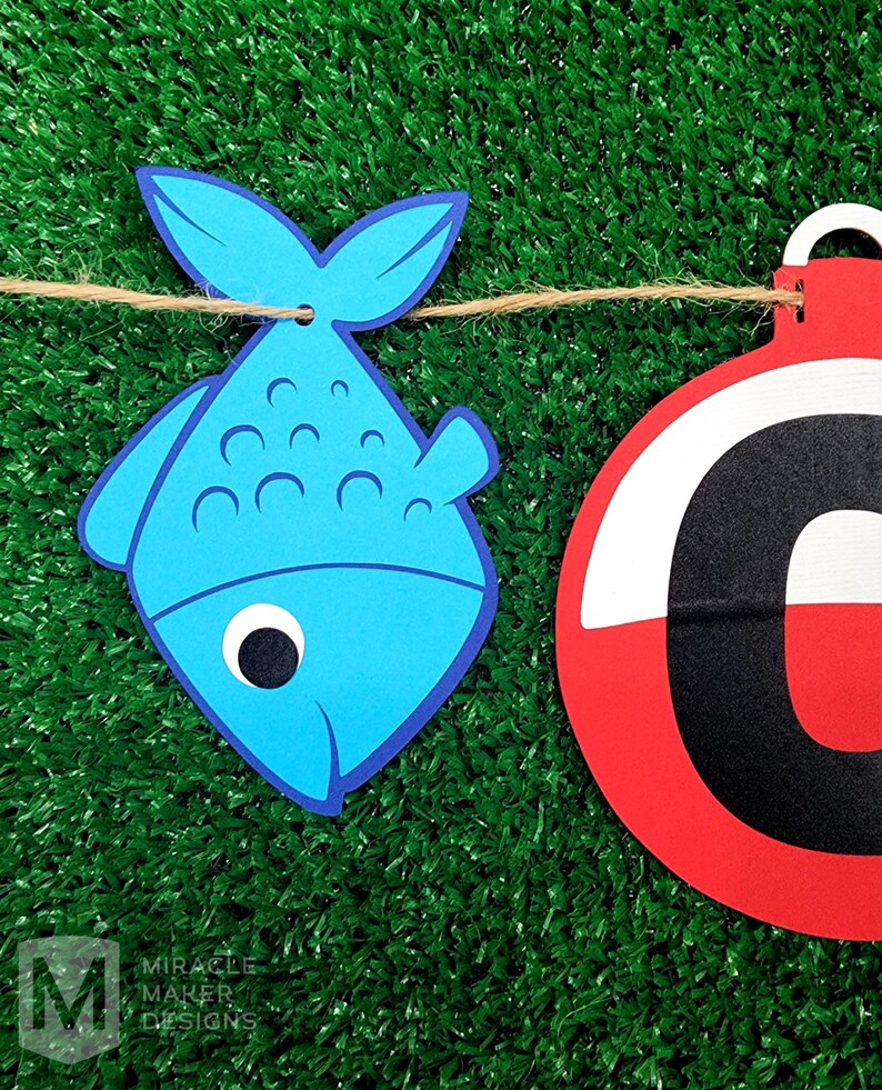 K&ouml;nnte beinhalten: Ein blauer Fisch und eine rot-wei&szlig;e Boje mit einem schwarzen Buchstaben C, die an einer Schnur h&auml;ngen. Der Hintergrund ist gr&uuml;nes Kunstgras.
