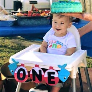 K&ouml;nnte beinhalten: Ein kleiner Junge in einem wei&szlig;en Shirt mit der Aufschrift "Fish All Day One" sitzt in einem Hochstuhl mit einem blauen und roten "One"-Banner. Das Baby tr&auml;gt schwarze Schuhe und hat einen blauen und gr&uuml;nen Kuchen auf dem Kopf.