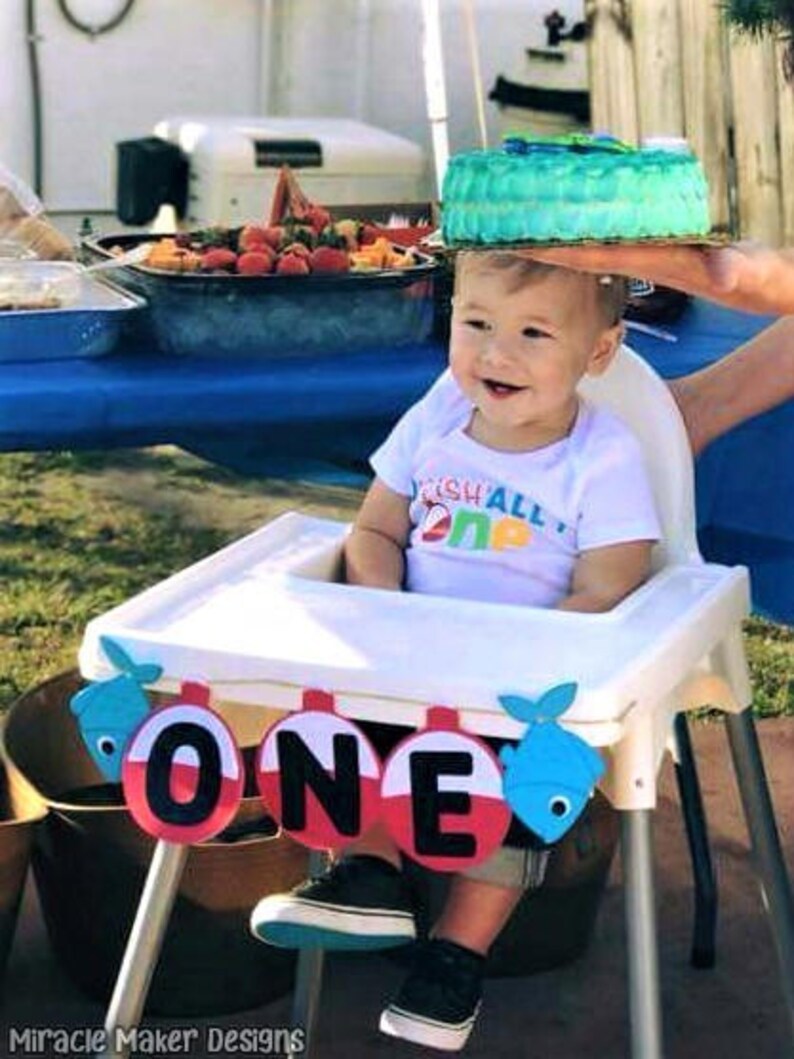 K&ouml;nnte beinhalten: Ein kleiner Junge in einem wei&szlig;en Shirt mit der Aufschrift "Fish All Day One" sitzt in einem Hochstuhl mit einem blauen und roten "One"-Banner. Das Baby tr&auml;gt schwarze Schuhe und hat einen blauen und gr&uuml;nen Kuchen auf dem Kopf.