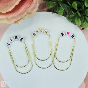 May include: Three gold-colored paper clips with white letter beads spelling "VIBE", "RKH", and "K♡R". The heart is pink. The paper clips are arranged on a white surface with a pink and green background.
