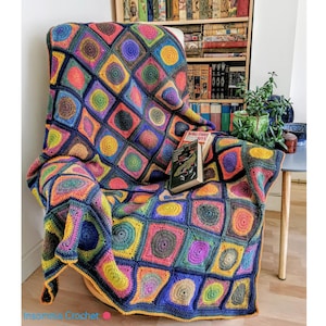 Op de afbeelding: Een kleurrijk gehaakt deken met een geometrisch patroon van vierkanten en cirkels in tinten blauw, groen, geel, rood en paars. Het deken is over een stoel in een woonkamer gelegd.