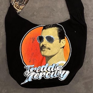 May include: Black shoulder bag featuring a graphic of Freddie Mercury in sunglasses against an orange circle. The bag has a paisley patterned strap and the words "Freddie Mercury" in a stylized font.