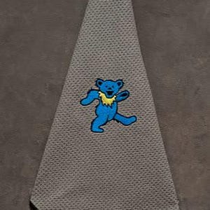May include: A triangular gray golf towel with a textured surface and a black carabiner clip at the top. The center of the towel features an embroidered graphic of a blue dancing bear with a yellow ruff around its neck, depicted in a mid-stride pose.