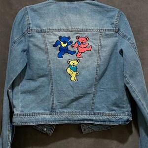 May include: Light blue denim jacket with three colorful dancing bear patches on the back. The patches feature bears in blue, red, and yellow with black outlines. The jacket has a classic collar, button closures, and pockets.