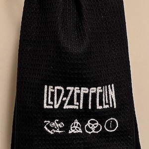 May include: Black waffle-weave towel with the Led Zeppelin band name and symbols embroidered in white. The towel is hanging from a silver ring. The towel is a dark, solid color.