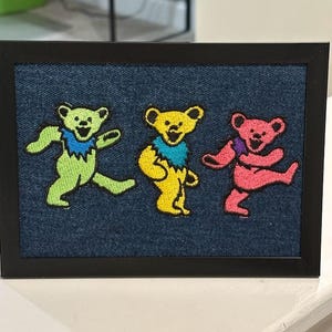 May include: A rectangular black frame containing a dark blue denim fabric background. Embroidered on the fabric are three dancing bears in a row: a light green bear with a blue collar, a yellow bear with a blue collar, and a pink bear.