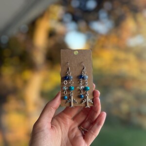 May include: Silver airplane earrings with blue and teal beads. The earrings are attached to a small, brown cardboard display card. The background is a blurred view of fall foliage.
