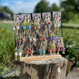 May include: A display of handmade macrame plant hangers with small terracotta pots and succulents. The hangers are in various colors, including red, orange, yellow, green, blue, purple, and white. The hangers are displayed on a wooden stand.