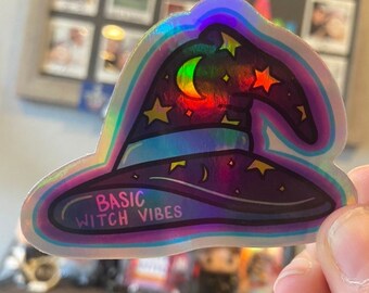 May include: A holographic sticker featuring a witch's hat, stars and a crescent moon. The sticker has the text "BASIC WITCH VIBES" on it.