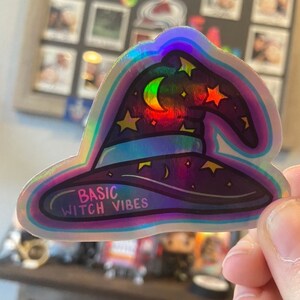 Pode incluir: Um adesivo holográfico com um chapéu de bruxa, estrelas e uma lua crescente. O adesivo tem o texto "BASIC WITCH VIBES".
