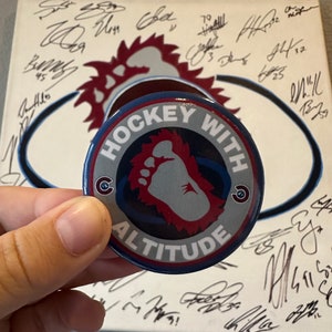 May include: A blue and red button with the text "Hockey With Altitude" and a stylized footprint graphic. The button is being held by a hand in front of a white surface with black signatures.