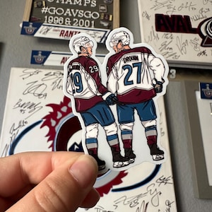 May include: A sticker featuring two hockey players wearing burgundy and blue jerseys with the numbers 29 and 27. The players are standing side-by-side, with the player on the left holding a hockey stick. The sticker is on a white background.