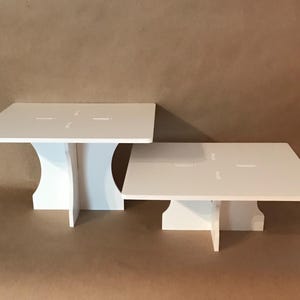 May include: Two white square cake stands with a simple design. The stands are made of sturdy cardboard and have a white finish. The larger stand is 12 inches wide and the smaller stand is 10 inches wide.