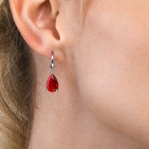 May include: A silver-toned earring with a teardrop-shaped, vibrant red gemstone. The earring is suspended from a small hook. The image is a close-up, highlighting the jewelry's color and design. The earring is a piece of fashion jewelry.