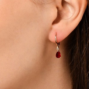 May include: A close-up of an ear with a gold earring featuring a teardrop-shaped, deep red gemstone and a small diamond. The leverback earring is gold with a polished finish. The earring is dangling.
