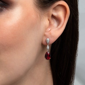 Könnte beinhalten: Eine Frau mit langen braunen Haaren trägt ein Paar Ohrringe mit roten birnenförmigen Edelsteinen und silbernen Akzenten.