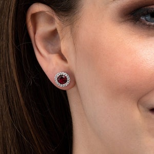 Puede incluir: Primer plano de una mujer con pendientes. Cada pendiente presenta una gema redonda de color rojo intenso, rodeada de piedras más pequeñas y transparentes. Los pendientes están engastados en metal plateado. La mujer sonríe.