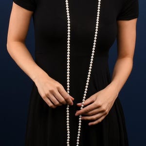 Puede incluir: Un collar largo de perlas blancas cuelga sobre un vestido negro. El collar está compuesto por muchas perlas pequeñas y redondas. El fondo es azul oscuro con un patrón floral.