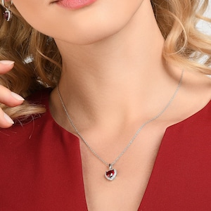 Può includere: Una donna che indossa un vestito rosso con una collana d'argento e orecchini con pietre rosse a forma di cuore. La collana e gli orecchini sono entrambi d'argento e hanno un design delicato.