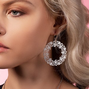 Może przedstawiać: Kobieta z blond włosami w czarnym topie i srebrnymi kolczykami w kształcie koła z delikatnym wzorem kwiatowym.