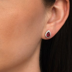 Könnte beinhalten: Eine Frau trägt ein Paar kleine, tropfenförmige Ohrringe mit einem roten Edelstein, der von einem Heiligenschein aus kleineren weißen Edelsteinen umgeben ist.