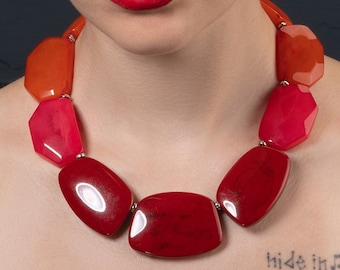 Puede incluir: Una mujer con el pelo corto castaño lleva un collar llamativo de color rojo, naranja y rosa. El collar está hecho de formas geométricas grandes y gruesas. La mujer mira directamente a la cámara.