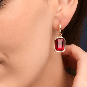 以下が含まれることがあります： 鮮やかな赤い宝石が付いた、金色の長方形のドロップイヤリング。イヤリングは金のフレームにセットされ、小さなフープから吊り下げられています。宝石は深く豊かな赤色で、エレガントさを添えています。