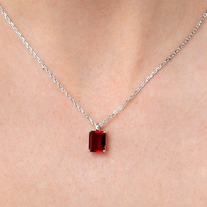 Pode incluir: Um colar de corrente de prata com um pequeno pingente quadrado de pedra vermelha.