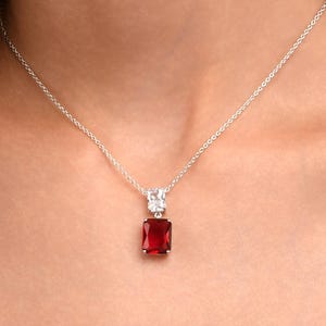 Puede incluir: Un collar de plata con un colgante rectangular de piedra preciosa rojo intenso. El colgante está adornado con piedras más pequeñas y transparentes. Se ve un pendiente a juego. La joya está sobre un fondo oscuro.