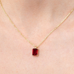 Puede incluir: Un collar de cadena de oro con un pequeño colgante cuadrado con una piedra preciosa roja.