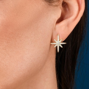 May include: A close-up of a woman's ear wearing a gold star-shaped earring with small clear stones. The earring is a simple and elegant design.