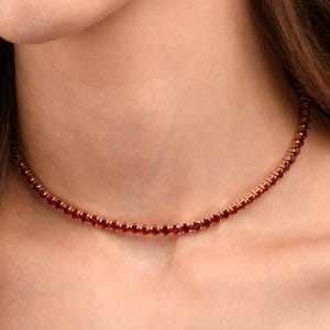 Peut inclure: Un collier doré orné d'une rangée de petites pierres précieuses rouges vives. Le collier est porté autour du cou, et l'image est un gros plan du bijou. Des boucles d'oreilles assorties en pierres précieuses rouges sont également visibles.